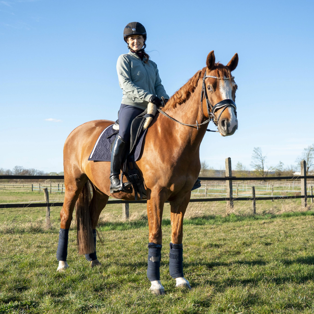 Blaue Dressur-Schabracke von ROSS KANI mit weißem Frühlingsband und Herz-Charm unter dem Sattel auf dem Pferd – stilvoller Look beim Reiten. Man sieht die Reiterin mit blauer Reithose auf dem Pferd.