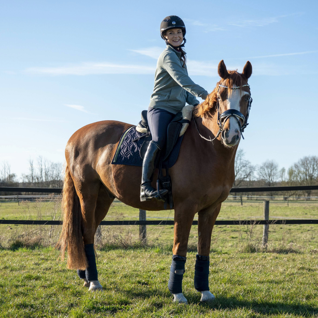Blaue Dressur-Schabracke von ROSS KANI mit grünem Frühlingsband und Gänseblümchen-Charm unter dem Sattel auf dem Pferd – stilvoller Look beim Reiten. Man sieht die Reiterin mit blauer Reithose auf dem Pferd.