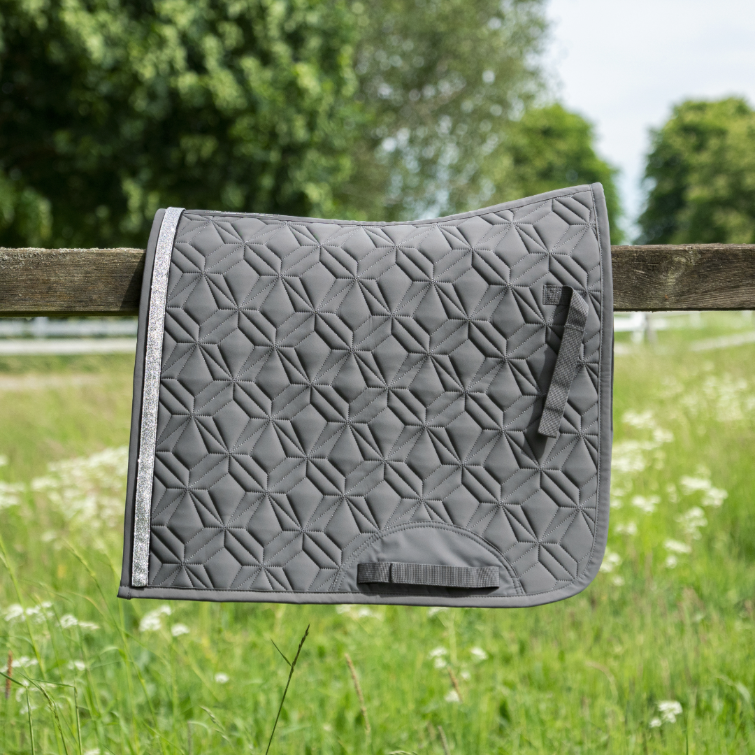 Graue ROSS KANI Dressur-Schabracke in Seitenansicht über einem Koppelzaun mit Wiese und Bäumen im Hintergrund– elegante Steppung und anatomische Passform.