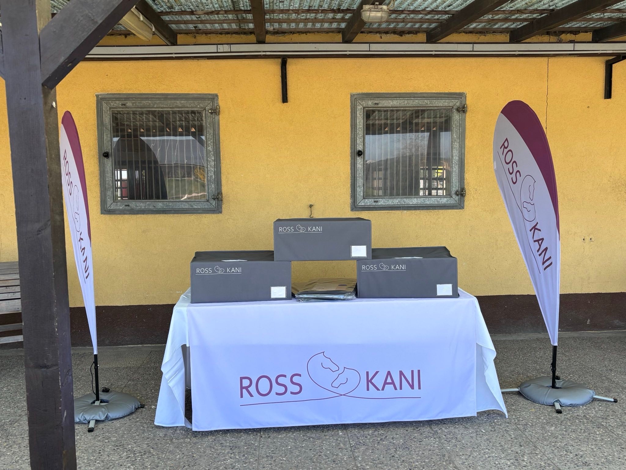 aufgebauter Verkaufsstand von ROSS KANI mit Beach-Flags und Aufbewahrungsboxen - Im Hintergrund sieht man den Pferdestall mit seinen Pferdeboxen