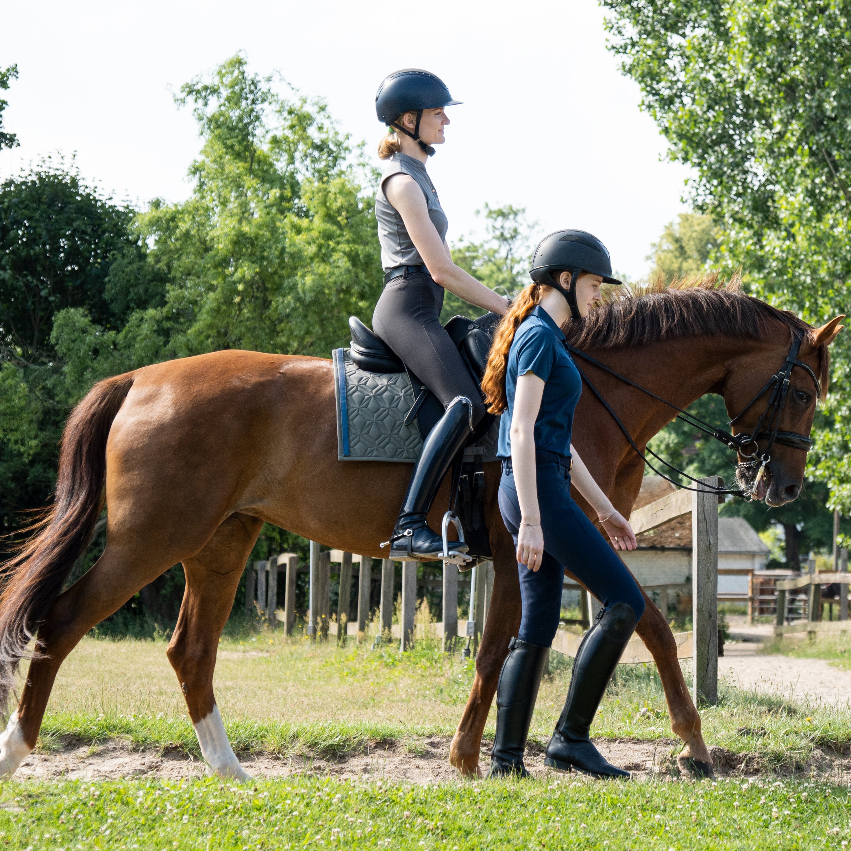 Graue Dressurschabracke von ROSS KANI mit blauem Glitzerband in sommerlicher Reitszene: Eine Reiterin sitzt im Schritt auf dem Pferd, während eine zweite Person in Reitkleidung daneben herläuft, warmes Licht und entspannte Atmosphäre.