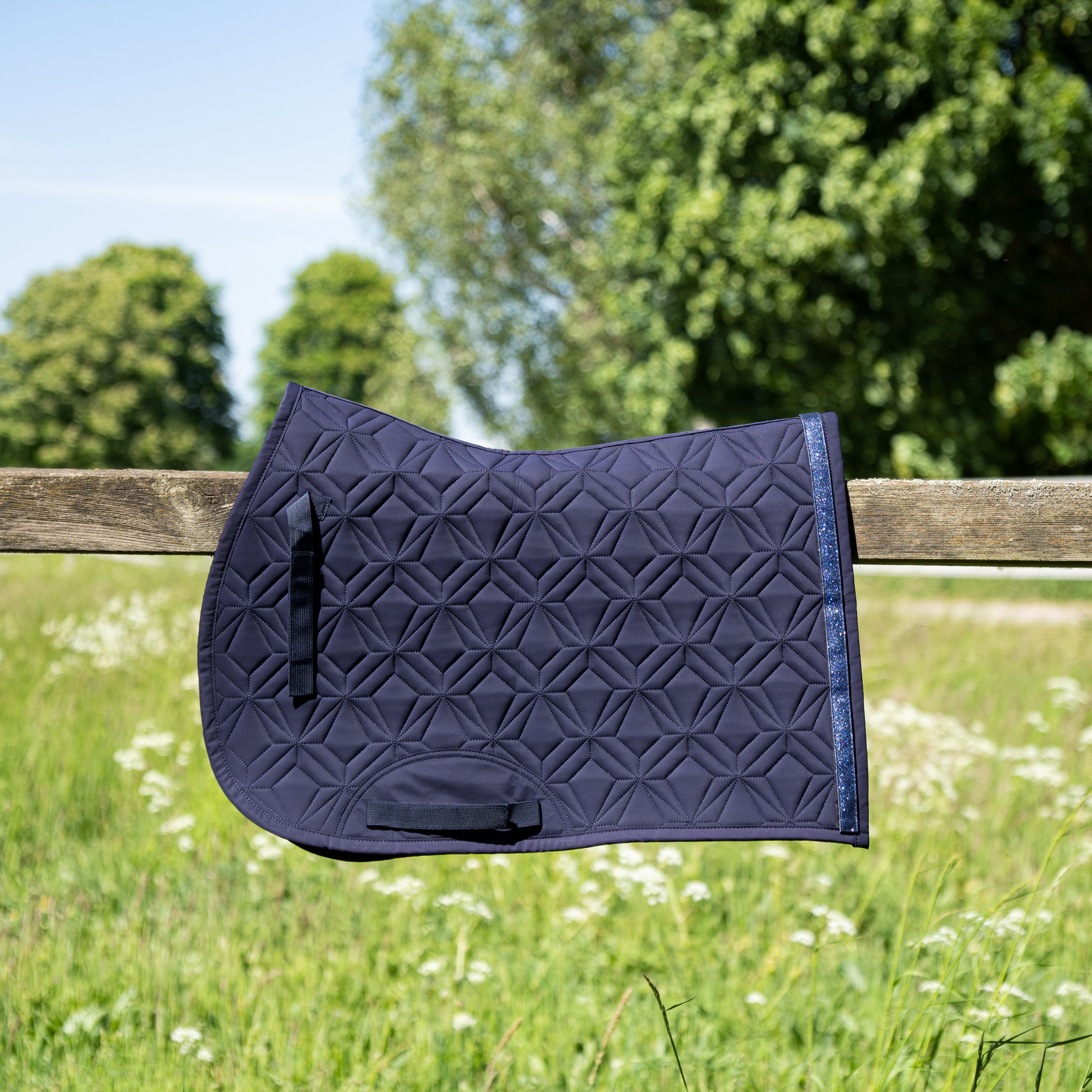 Blaue Springschabracke von ROSS KANI in Seitenansicht über einem Koppelzaun mit Wiese und Bäumen im Hintergrund– elegante Steppung und anatomische Passform.