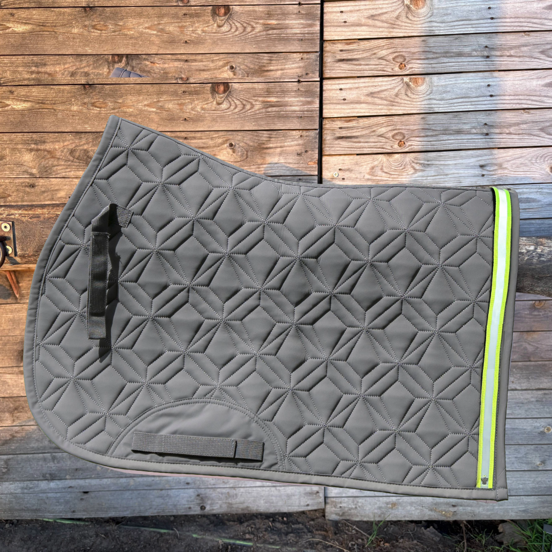 Graue Spring-Hauptschabracke von ROSS KANI hängt über einem Holzzaun. Es ist ein neonfarbenes Klettband mit grauem Reflektorstreifen und einem kleinen silbernen Anhänger in Engelform aufgeklettet. Im Hintergrund sieht man etwas sandigen Boden mit Grashalmen und eine Wand aus Holzbrettern.