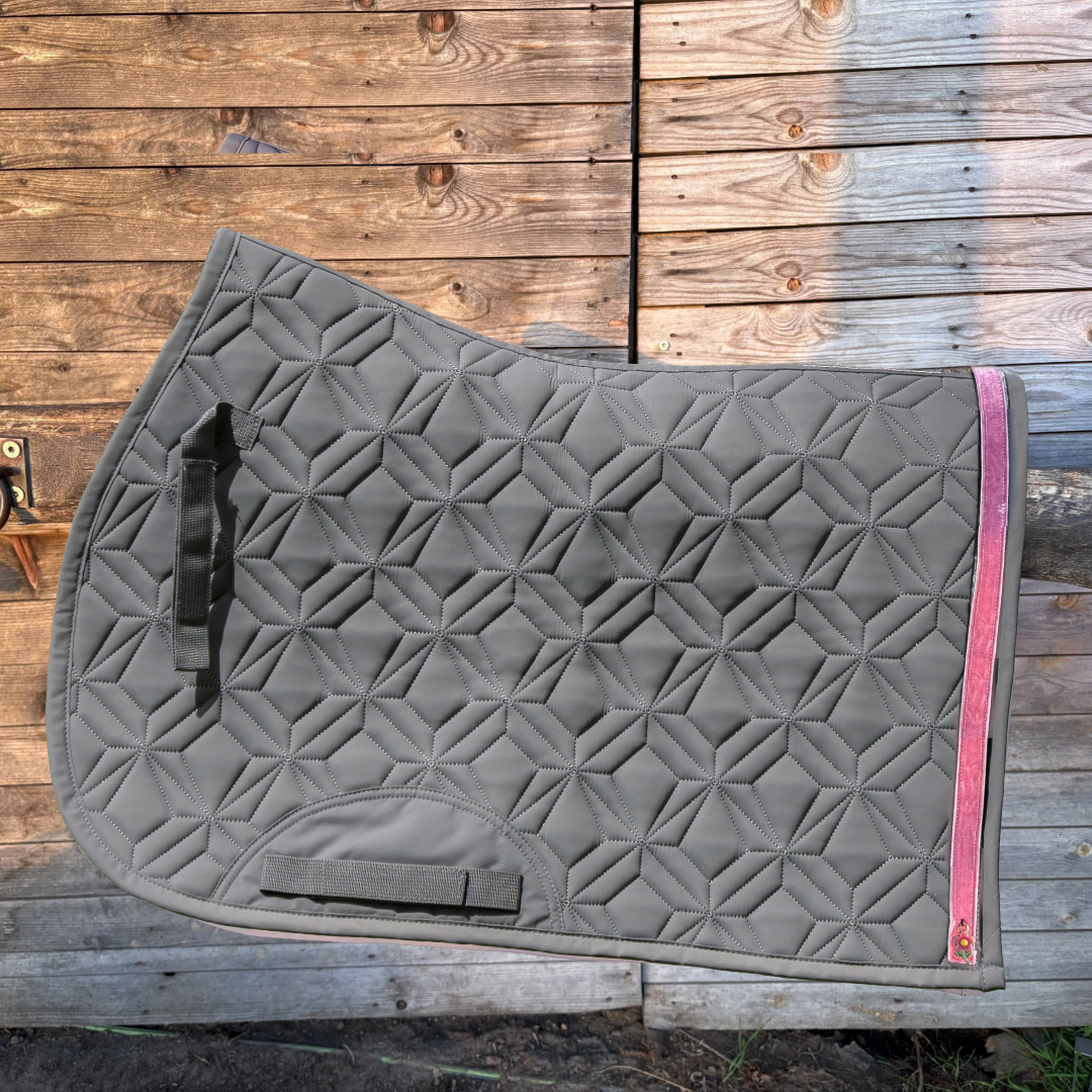 Graue Springschabracke von ROSS KANI mit rosa Frühlingsband und Blumen-Charm vor einer Holzwand.