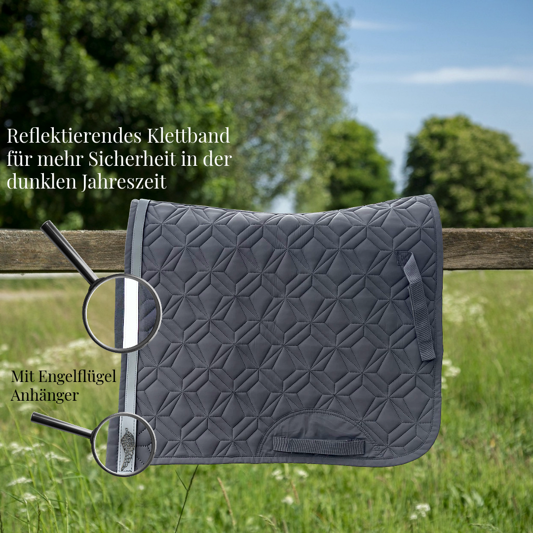 Grau, gesteppte Dressurschabracke von ROSS KANI hängt über einem Holzzaun auf einer Wiese. Ein reflektierendes Klettband mit Engelflügel-Charm ist am Rand befestigt und wird durch eine Lupe hervorgehoben. Text im Bild weist auf erhöhte Sicherheit in der dunklen Jahreszeit hin.