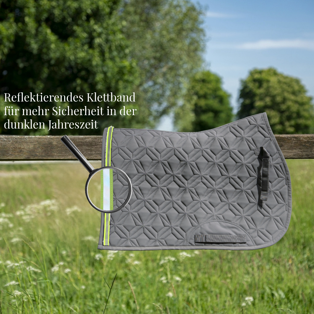 Graue, gesteppte Springschabracke von ROSS KANI hängt über einem Holzzaun auf einer Wiese. Ein reflektierendes Klettband in Neonfarbe ist am Rand befestigt und wird durch eine Lupe hervorgehoben. Text im Bild weist auf erhöhte Sicherheit in der dunklen Jahreszeit hin.