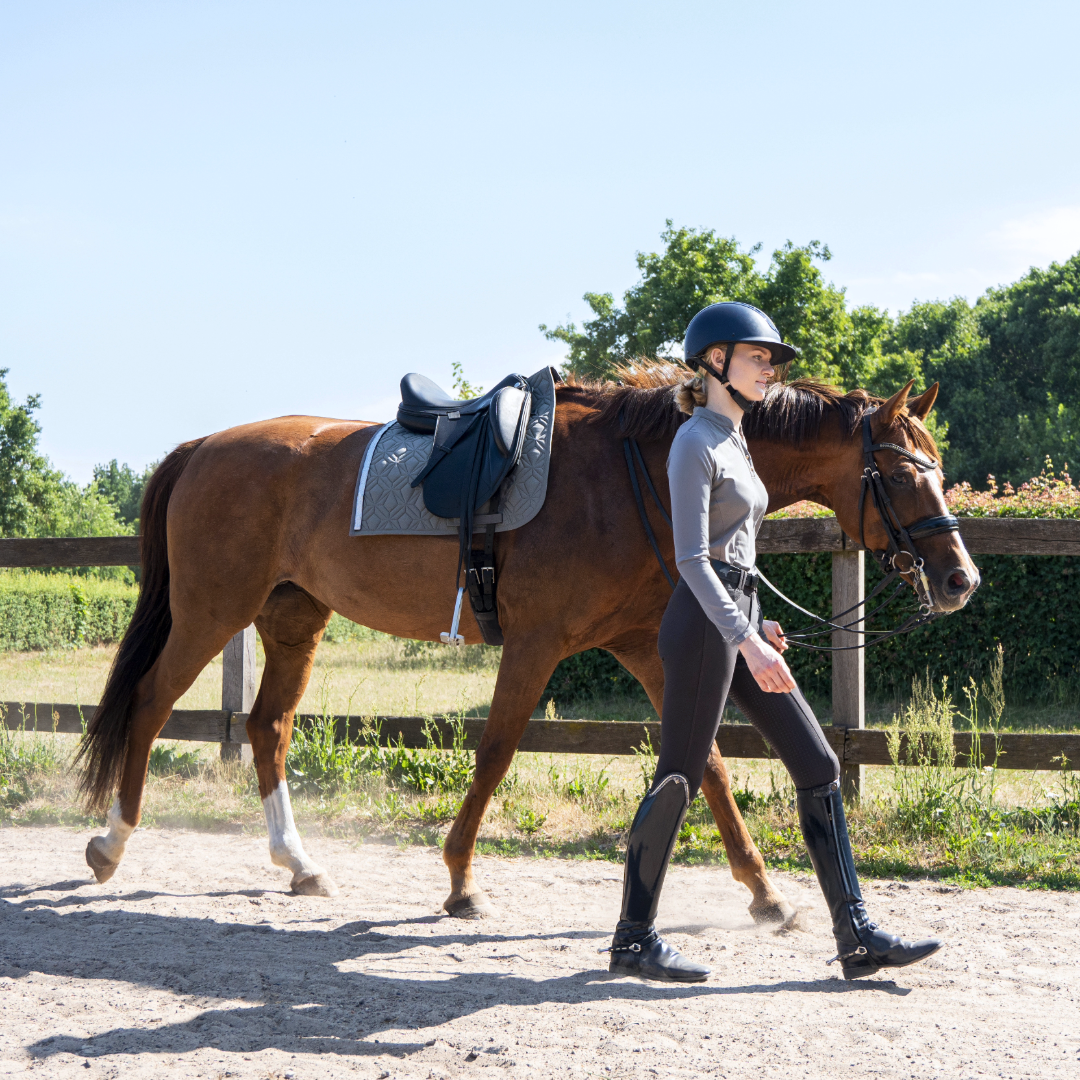 Reiterin führt ein Pferd vom Reitplatz. Das Pferd ist gesattelt und unter dem Sattel liegt die graue Dressurschabracke von ROSS KANI. Die Reiterin trägt Reithelm, Reitstiefel und enganliegende Reitbekleidung. Sonniger Tag mit Holzzaun und grünen Bäumen im Hintergrund.