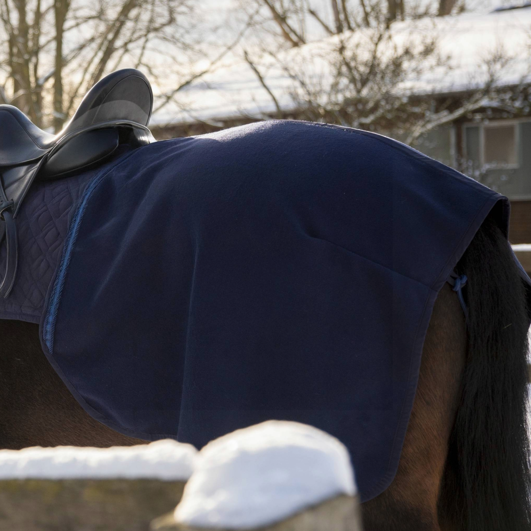 Die blaue Wollwalk-Ausreitdecke von ROSS KANI ist auf der blauen Hauptschabracke befestigt und liegt unter dem Sattel auf einem dunkelbraunen Pferd. Im Hintergrund sieht man Bäume und ein Gebäude im Schnee.