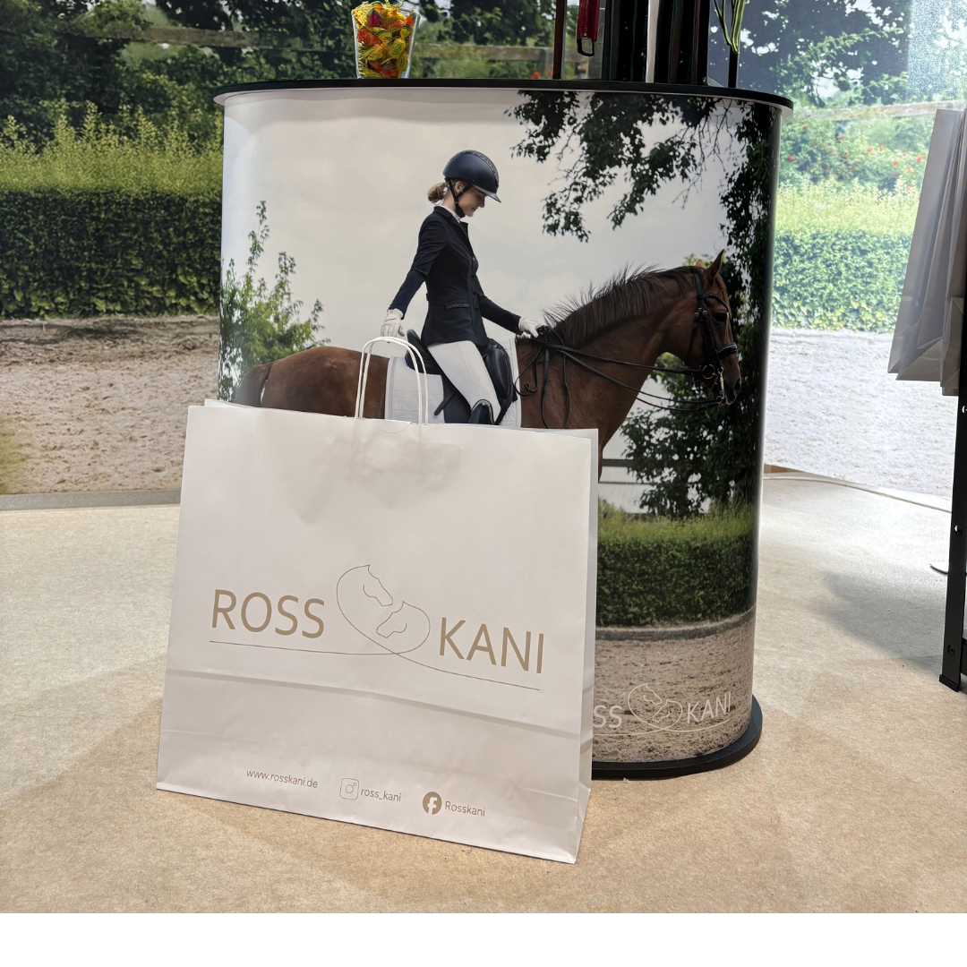 Counter auf der Messe mit der Einkaufstüte von ROSS KANI im Vordergrund. Auf dem Counter ist ein Pferd mit Reiterin zur Grußaufstellung in Seitenansicht. Die Reiterin trägt eine weiße Reithose mit schwarzer Turnierjacke und unter dem Sattel liegt das weiße Dressur-Cover von ROSS KANI.