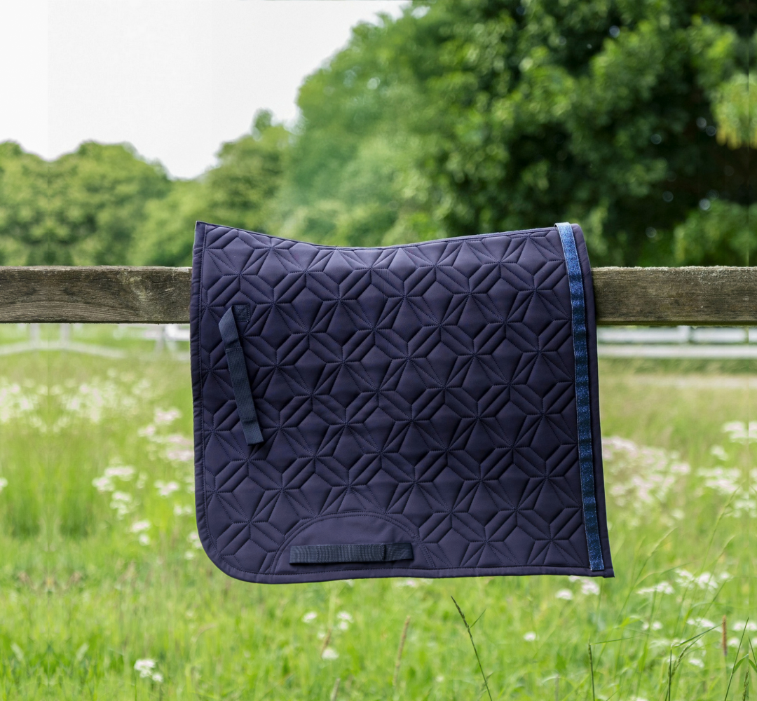 Blaue ROSS KANI Dressur-Schabracke in Seitenansicht über einem Koppelzaun mit Wiese und Bäumen im Hintergrund– elegante Steppung und anatomische Passform.