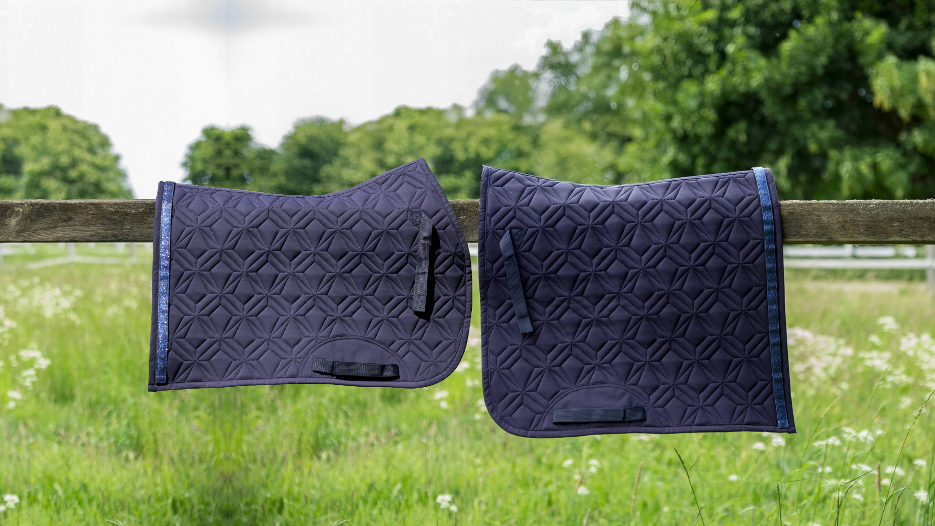 Blaue Dressurschabracke und blaue Springschabracke von ROSS KANI in Seitenansicht über einem Koppelzaun mit Wiese und Bäumen im Hintergrund– elegante Steppung und anatomische Passform.