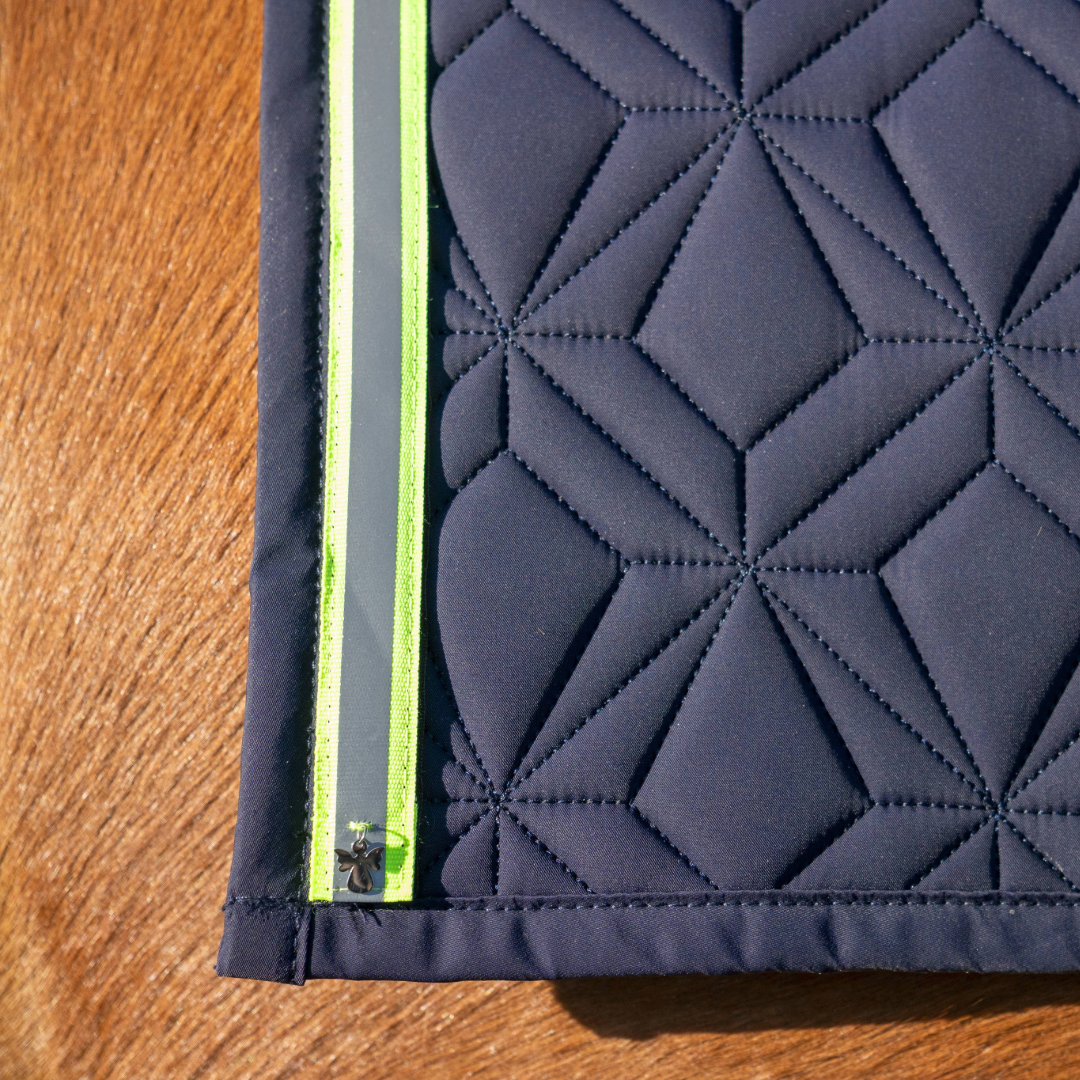 Blaue Schabracke von ROSS KANI mit neonfarbenen Reflektorband mit Schutzengel-Charm auf dem Pferd – sicherer Look beim Reiten. 