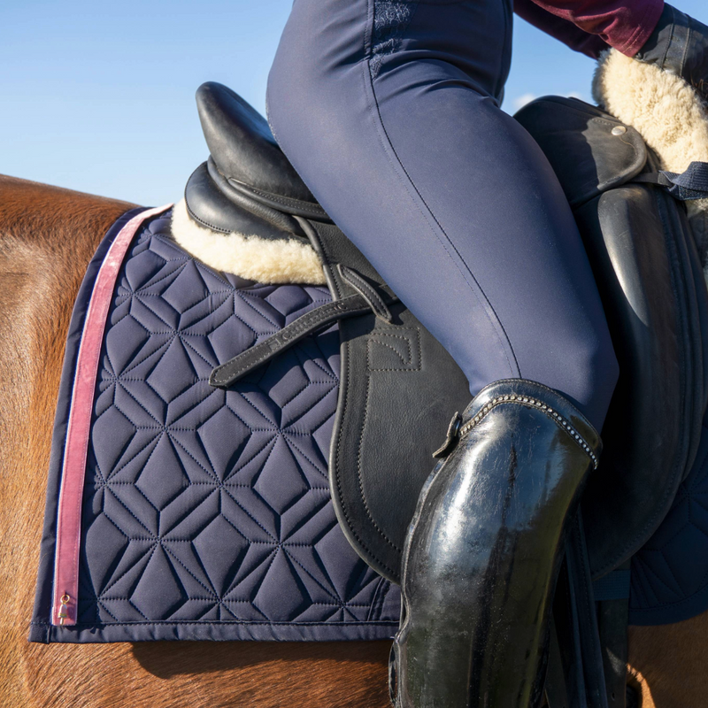 Blaue Dressur-Schabracke von ROSS KANI mit rosafarbenem Frühlingsband unter dem Sattel auf dem Pferd – stilvoller Look beim Reiten. Man sieht eine blaue Reithose und einen schwarzen Stiefel in Seitenansicht.
