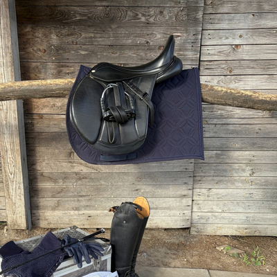 Dunkelblaue ROSS KANI Classic Spring-Schabracke mit schwarzem Springsattel und Reitzubehör vor einer Holzwand