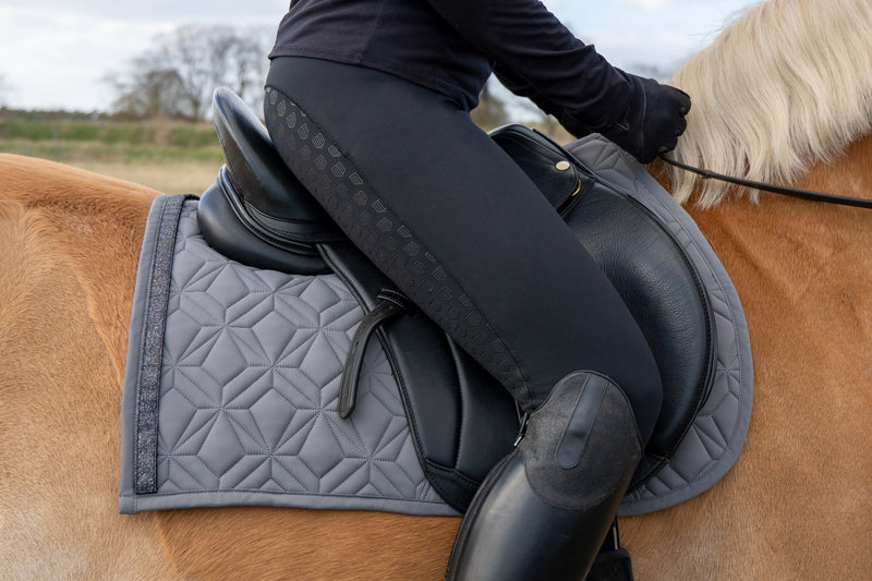 Graue Springschabracke von ROSS KANI mit schwarzem Glitzerband unter dem Sattel auf dem Pferd – stilvoller Look beim Reiten. Man sieht eine schwarze Reithose und einen schwarzen Stiefel in Seitenansicht.