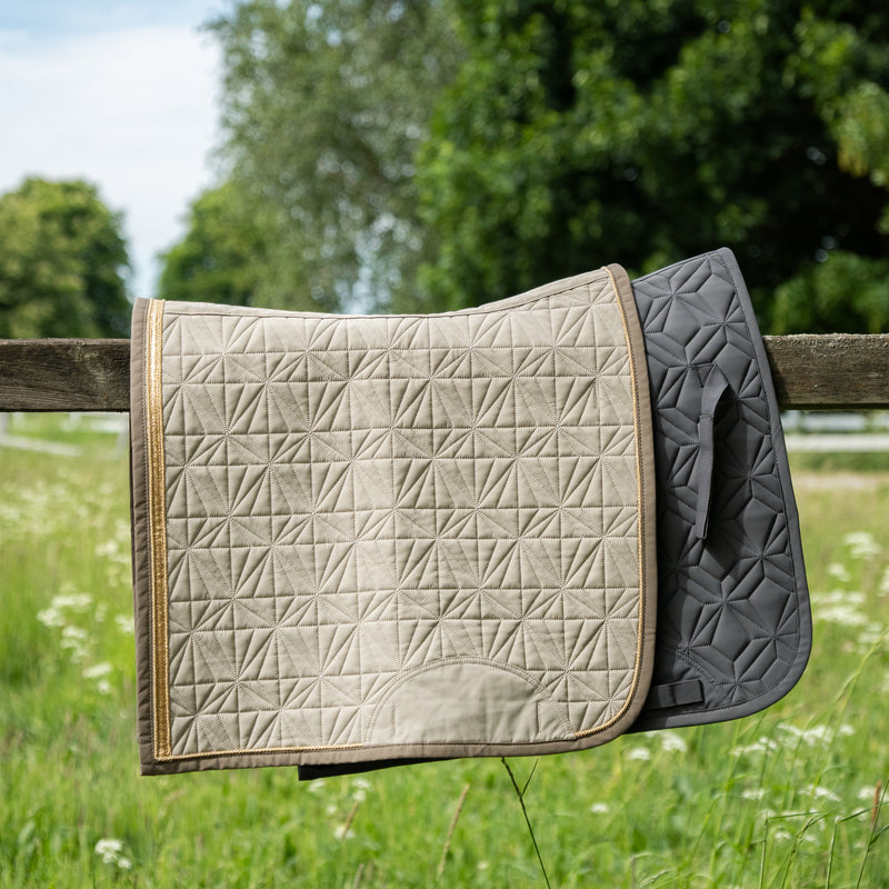 Graue Dressur-Schabracke und Dressur Cover beige von ROSS KANI in Seitenansicht über einem Koppelzaun mit Wiese und Bäumen im Hintergrund – passendes Set für jeden Ritt.