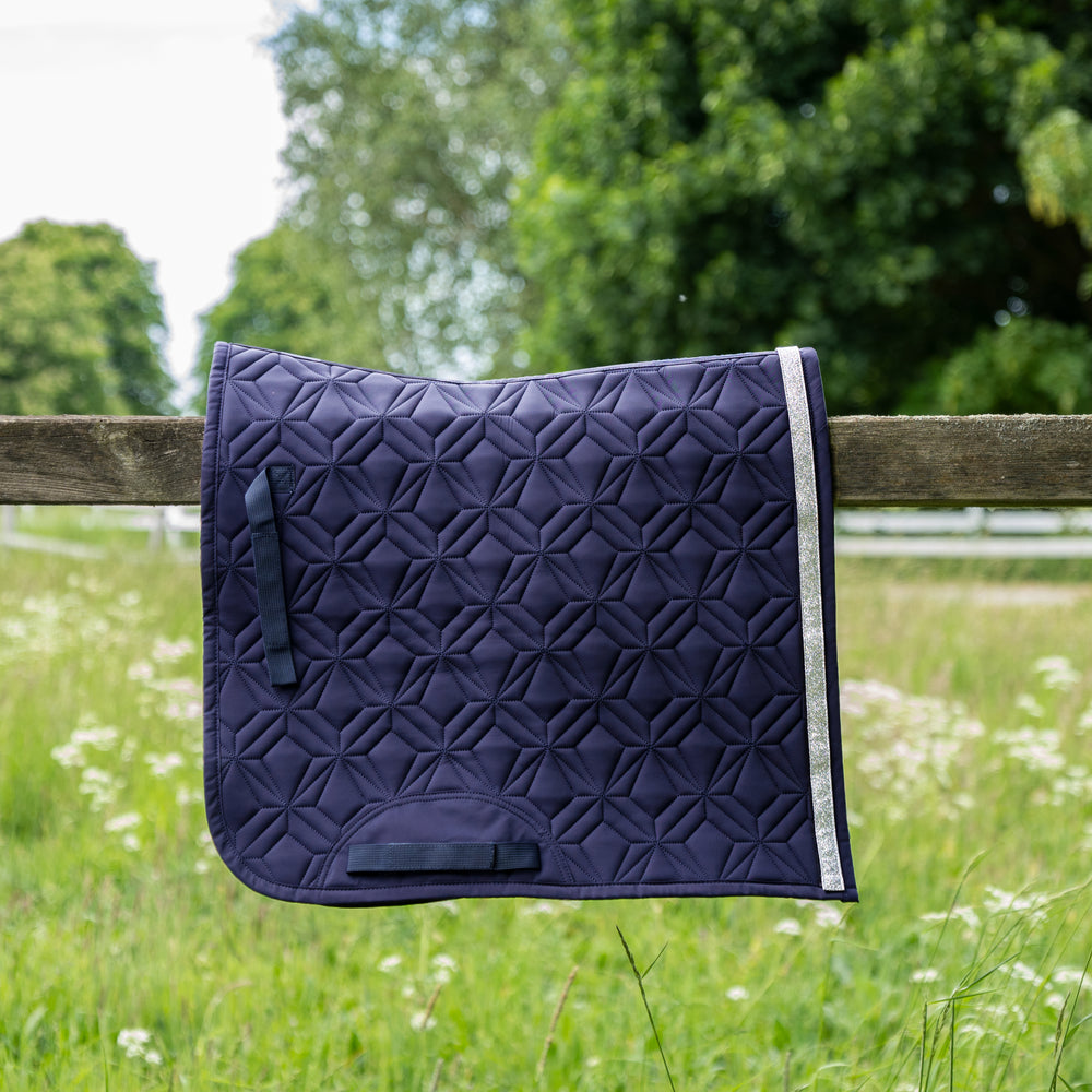 Blaue ROSS KANI Dressur-Schabracke mit silbernem Glitzerband in Seitenansicht über einem Koppelzaun mit Wiese und Bäumen im Hintergrund– elegante Steppung und anatomische Passform.
