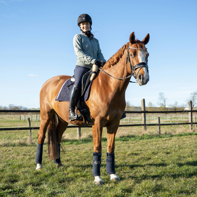 Blaue Dressur-Schabracke von ROSS KANI mit weißem Frühlingsband und Herz-Charm unter dem Sattel auf dem Pferd – stilvoller Look beim Reiten. Man sieht die Reiterin mit blauer Reithose auf dem Pferd.