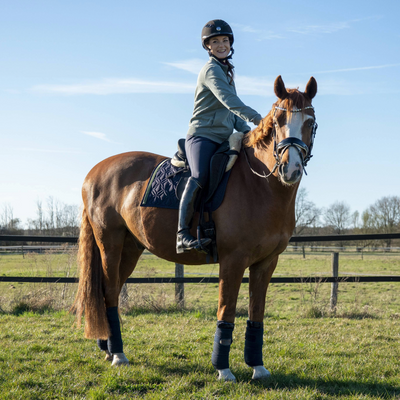 Blaue Dressur-Schabracke von ROSS KANI mit grünem Frühlingsband und Gänseblümchen-Charm unter dem Sattel auf dem Pferd – stilvoller Look beim Reiten. Man sieht die Reiterin mit blauer Reithose auf dem Pferd.