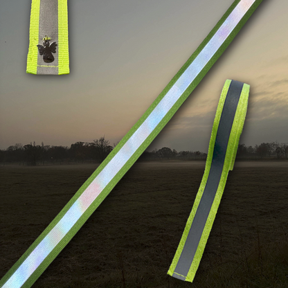 Reflektierende Bänder in Neonfarbe mit Schutzengel-Charm von ROSS KANI vor einem nebelig dunklem Hintergrund. Die schmalen Bänder zeigen eine helle, reflektierende Mittelstreifenfläche mit neonfarbenen Kanten.