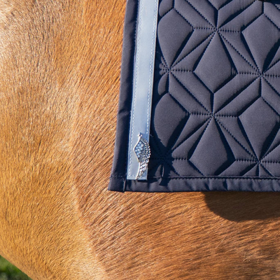 Detailansicht der blauen Hauptschabracke mit grauem Reflektorband und silbernem Flügelanhänger. Die Schabracke liegt auf einem fuchsfarbenem Pferd.