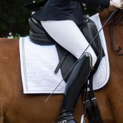 Weißes Dressur-Cover von ROSS KANI unter dem Sattel auf dem Pferd – stilvoller Look beim Turnier. Man sieht eine weiße Reithose und einen schwarzen Stiefel in Seitenansicht.