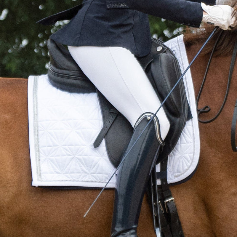 Weißes Dressur-Cover von ROSS KANI unter dem Sattel auf dem Pferd – stilvoller Look beim Turnier. Man sieht eine weiße Reithose und einen schwarzen Stiefel in Seitenansicht.