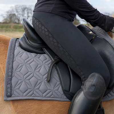 Graue Springschabracke von ROSS KANI mit schwarzem Glitzerband unter dem Sattel auf dem Pferd – stilvoller Look beim Reiten. Man sieht eine schwarze Reithose und einen schwarzen Stiefel in Seitenansicht.