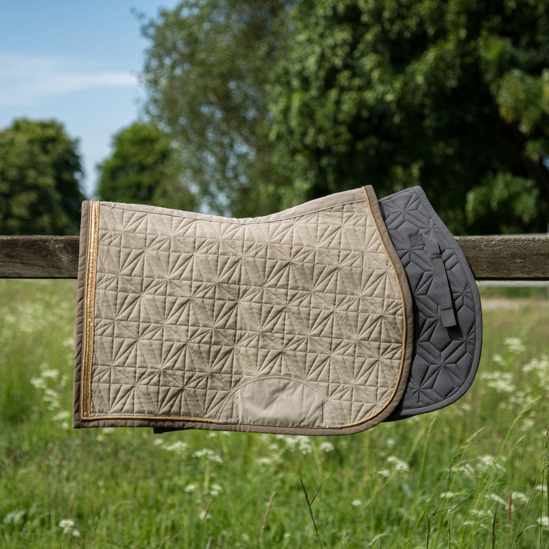 Graue Springschabracke und Cover Springen beige von ROSS KANI in Seitenansicht über einem Koppelzaun mit Wiese und Bäumen im Hintergrund – passendes Set für jeden Ritt.
