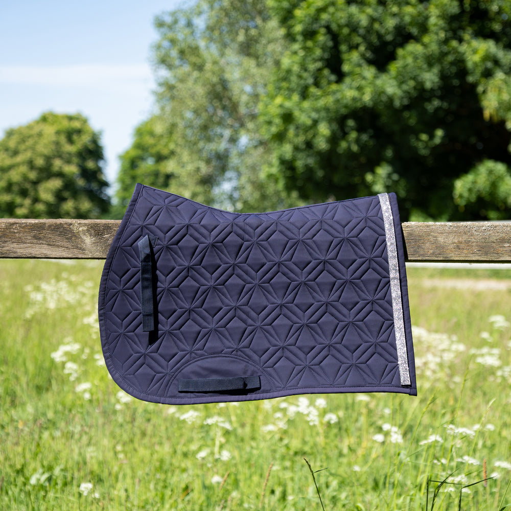 Blaue ROSS KANI Springschabracke mit silbernem Glitzerband in Seitenansicht über einem Koppelzaun mit Wiese und Bäumen im Hintergrund– elegante Steppung und anatomische Passform.