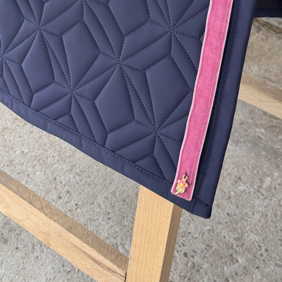 Detailansicht der blauen Dressur-Hauptschabracke von ROSS KANI mit aufgeklettetem Samtband in rosa mit kleinem Anhänger in Blumenform mit rosa Blüte. Im Hintergrund ist Steinboden und der untere Teil eines Holzbocks zu sehen.