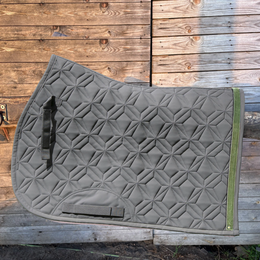 Graue Springschabracke von ROSS KANI mit grünem Frühlingsband und Gänseblümchen-Charm vor einer Holzwand.