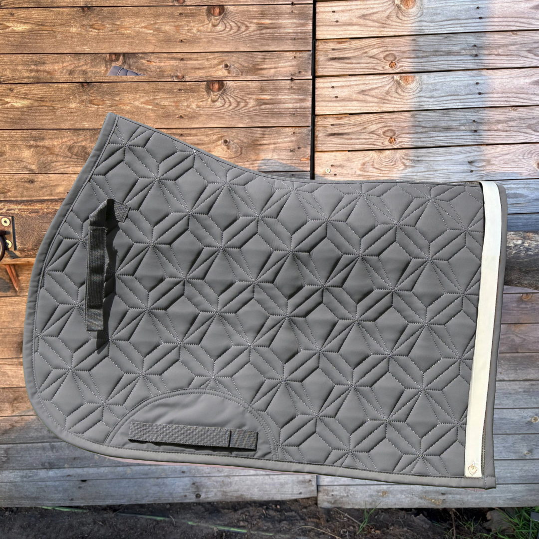 Graue Springschabracke von ROSS KANI mit weißem Frühlingsband und rosé Herz-Charm vor einem Holzzaun.

