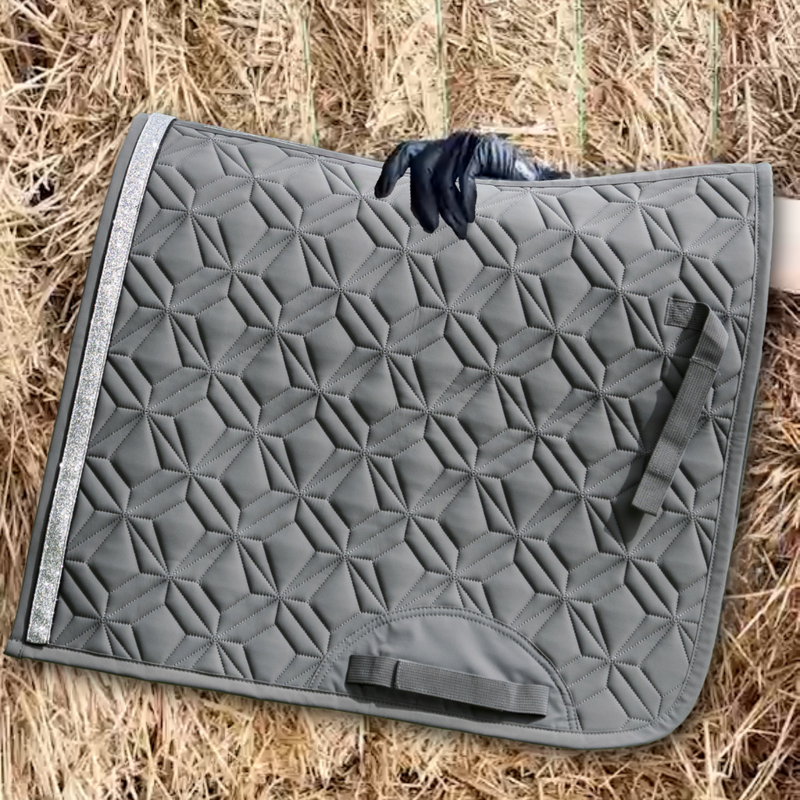 Graue Dressurschabracke von ROSS KANI wird vor einen Strohhintergrund gehalten.