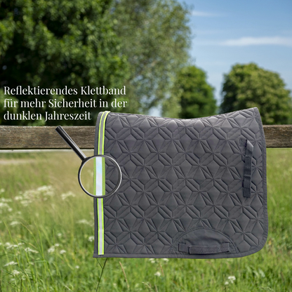 Graue, gesteppte Dressurschabracke von ROSS KANI hängt über einem Holzzaun auf einer Wiese. Ein reflektierendes Klettband in Neonfarbe ist am Rand befestigt und wird durch eine Lupe hervorgehoben. Text im Bild weist auf erhöhte Sicherheit in der dunklen Jahreszeit hin.
