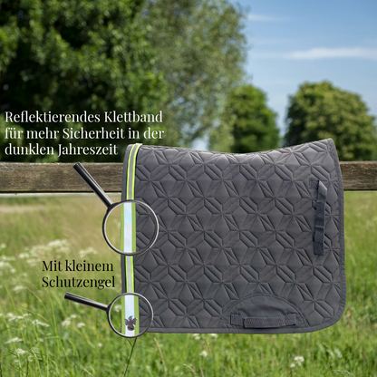 Graue, gesteppte Dressurschabracke von ROSS KANI hängt über einem Holzzaun auf einer Wiese. Ein reflektierendes Klettband in Neonfarbe mit Schutzengel-Charm ist am Rand befestigt und wird durch eine Lupe hervorgehoben. Text im Bild weist auf erhöhte Sicherheit in der dunklen Jahreszeit hin.