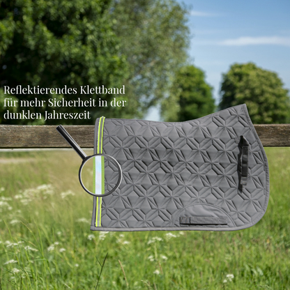 Graue, gesteppte Springschabracke von ROSS KANI hängt über einem Holzzaun auf einer Wiese. Ein reflektierendes Klettband in Neonfarbe ist am Rand befestigt und wird durch eine Lupe hervorgehoben. Text im Bild weist auf erhöhte Sicherheit in der dunklen Jahreszeit hin.