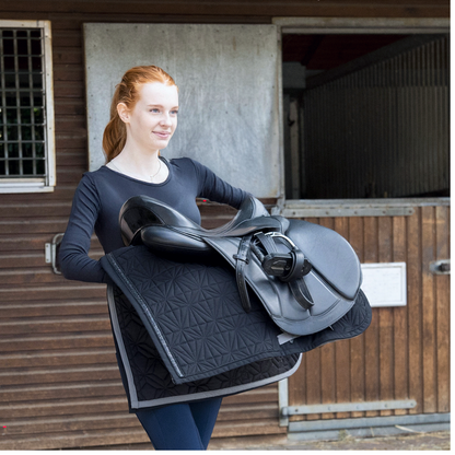 Reiterin in Reitbekleidung trägt ROSS KANI Schabracken-Cover in Schwarz mit schwarzem Vielseitigkeitssattel vor einer Box – stilvolles Reitequipment für Springreiten und Alltag.