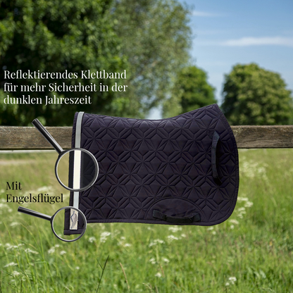 Blau, gesteppte Springschabracke von ROSS KANI hängt über einem Holzzaun auf einer Wiese. Ein reflektierendes Klettband mit Engelflügel-Charm ist am Rand befestigt und wird durch eine Lupe hervorgehoben. Text im Bild weist auf erhöhte Sicherheit in der dunklen Jahreszeit hin.