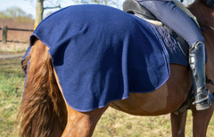 Blaue Ausreitdecke von ROSS KANI auf dem Pferd, kombiniert mit passender Dressurschabracke; Reiterin sitzt im Sattel auf einer Wiese mit wenig Bewuchs, Büsche im Hintergrund.