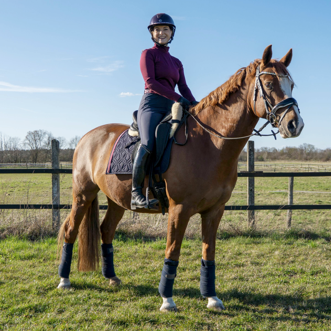 Blaue Dressur-Schabracke von ROSS KANI mit rosa Frühlingsband und Eis-Charm unter dem Sattel auf dem Pferd – stilvoller Look beim Reiten. Man sieht die Reiterin mit blauer Reithose auf dem Pferd.