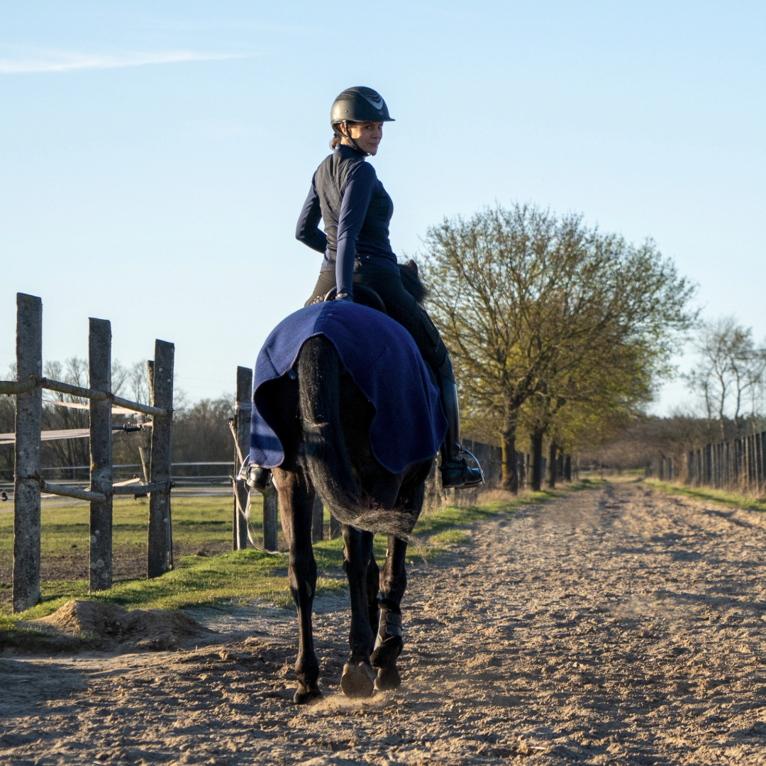 Gründerin Katrin sitzt auf einem Pferd und dreht sich nach hinten in die Kamera. Man sieht die dunkelblaue Ausreitdecke von ROSS KANI auf dem Pferd liegen, im Hintergrund Himmel und Bäume.
