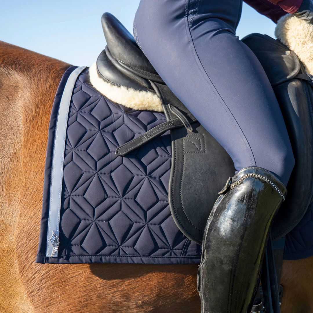 Blaue Dressur-Schabracke von ROSS KANI mit reflektierendem Klettband in Silber mit silbernem Anhänger in Flügelform. Die Schabracke liegt auf einem fuchsfarbenem Pferd, darauf liegt ein schwarzer Sattel. Es ist ein Stück Bein mit blauer Reithose und schwarzem Stiefel zu sehen.