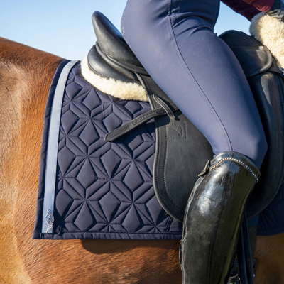 Blaue Dressur-Schabracke von ROSS KANI mit reflektierendem Klettband in Silber mit silbernem Anhänger in Flügelform. Die Schabracke liegt auf einem fuchsfarbenem Pferd, darauf liegt ein schwarzer Sattel. Es ist ein Stück Bein mit blauer Reithose und schwarzem Stiefel zu sehen.