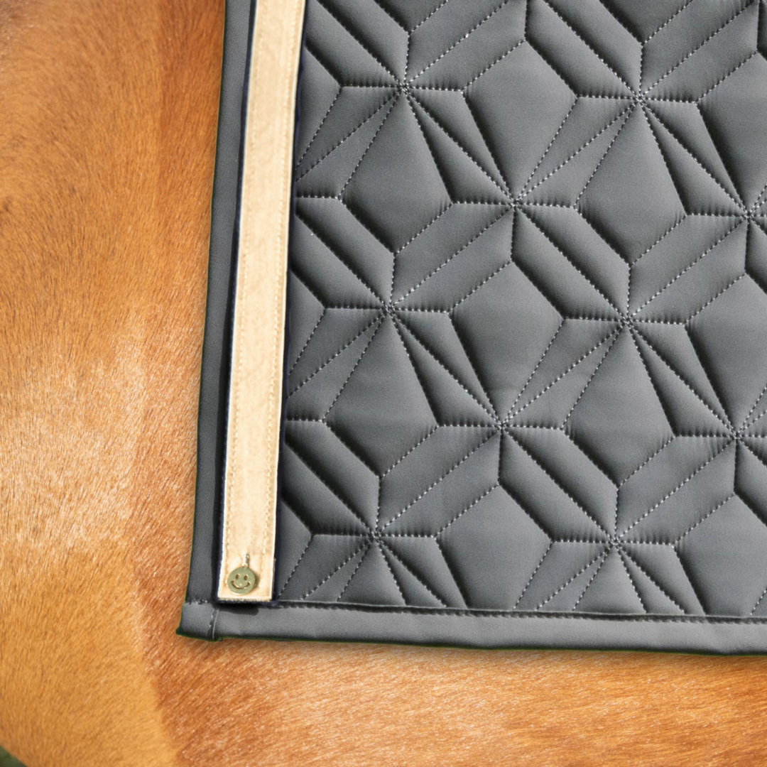 Graue Schabracke von ROSS KANI mit beigem Frühlingsband und Smiley-Charm unter dem Sattel auf dem Pferd – stilvoller Look beim Reiten. 