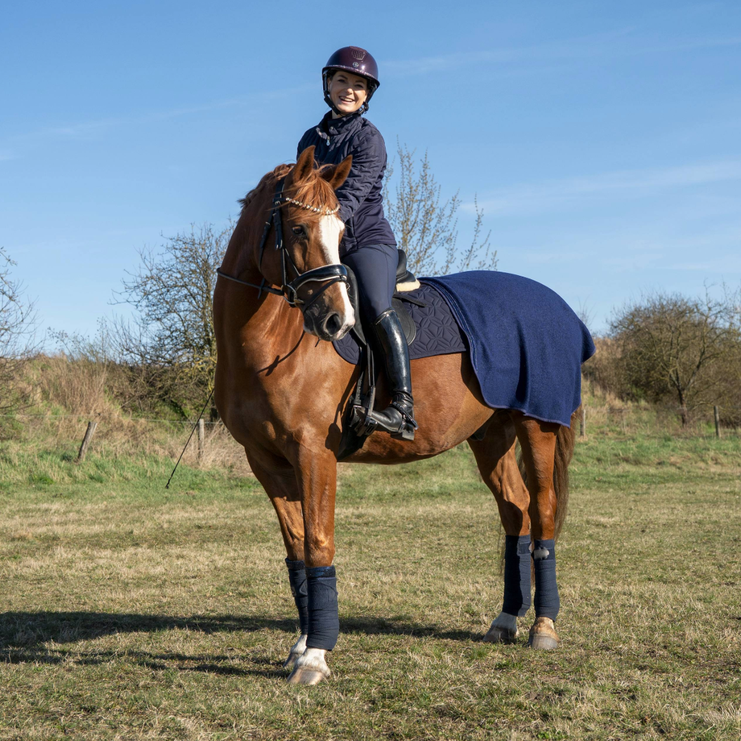 Blaue Ausreitdecke von ROSS KANI auf dem Pferd, kombiniert mit passender Dressurschabracke; Reiterin sitzt im Sattel auf einer Wiese mit wenig Bewuchs, Büsche im Hintergrund.
