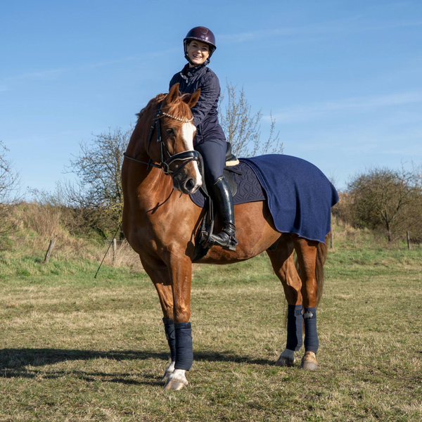 Blaue Ausreitdecke von ROSS KANI auf dem Pferd, kombiniert mit passender Dressurschabracke; Reiterin sitzt im Sattel auf einer Wiese mit wenig Bewuchs, Büsche im Hintergrund.