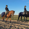 Video: Zwei Reiterinnen reiten in der Morgensonne an einer Koppel entlang. Beide haben die blaue Ausreitdecke von ROSS KANI an ihren Schabracken befestigt.  Die eine Reiterin an der blauen Dressurschabracke, die andere an der grauen.
